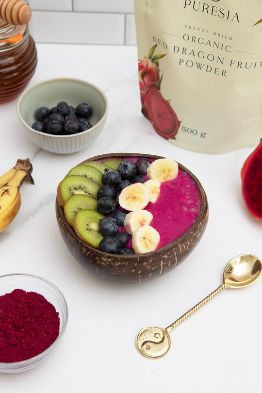 Coconut bowl with fruit and a packet of Puresia Organic Red Dragon Fruit Powder on a white surface.